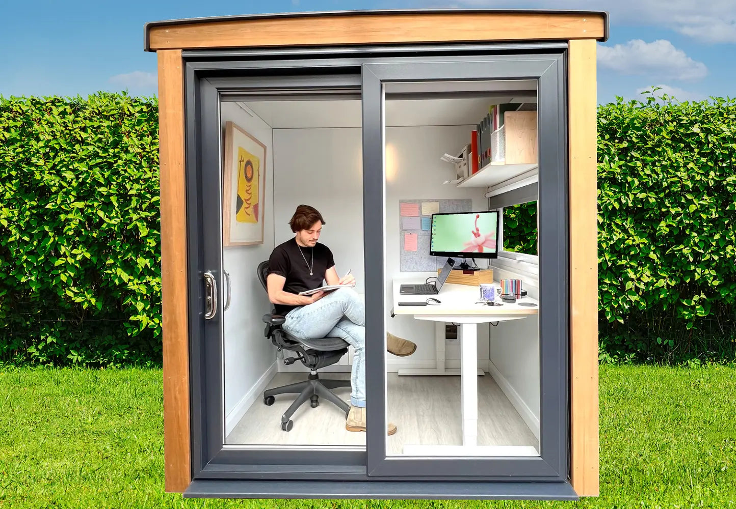 Garden Office 2A, Small office pod situated in a garden. with patio door and window on the right side. Cladding Western Red Cedar. Model sitting in the office pod in an executive chair, table and computer.